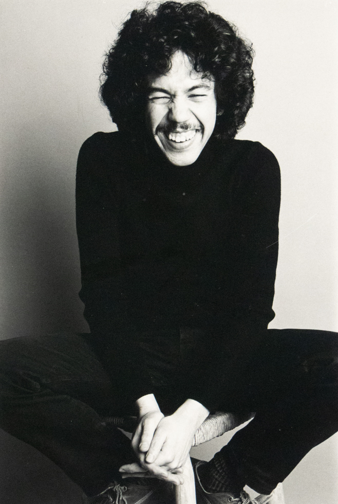 A black and white studio portrait captures a person with curly hair and a mustache, seated and smiling broadly, wearing a dark turtleneck and dark pants. They are looking directly at the viewer with visible joy.