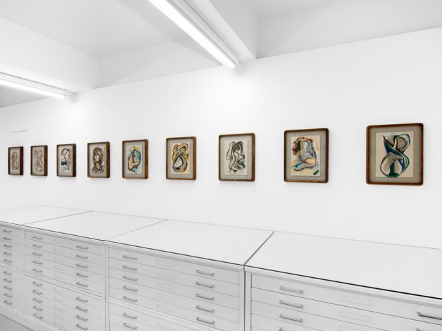 A series of abstract framed drawings hang on a white gallery wall under a black and white artist name and exhibition title.