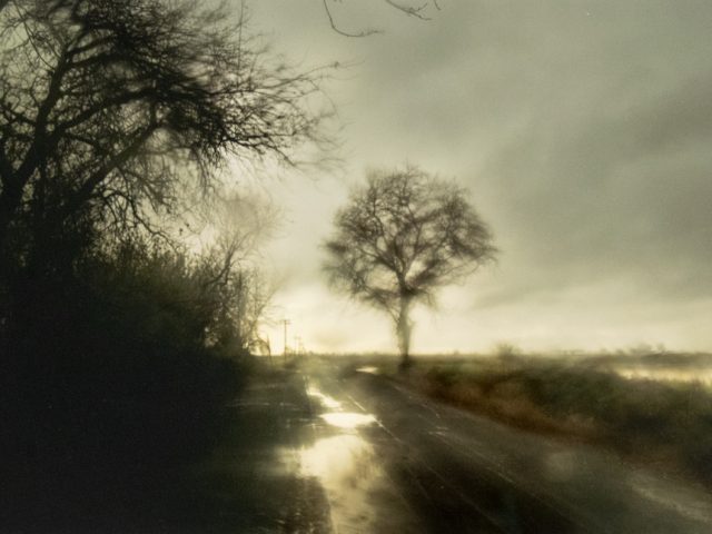 Blurry rural road in rain with bare trees and a moody, overcast sky.