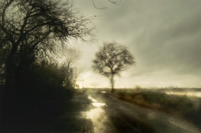 Blurry rural road in rain with bare trees and a moody, overcast sky.