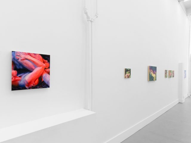 Colorful paintings hang on bright white gallery walls with grey floors.