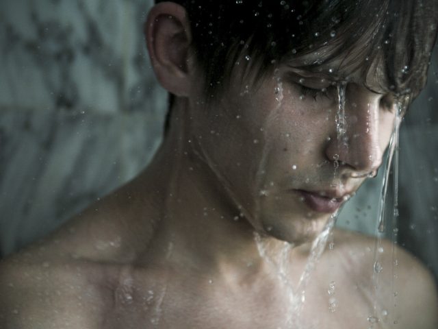 Shirtless young man with wet hair stands under shower water with eyes closed, appearing introspective against a marble background.