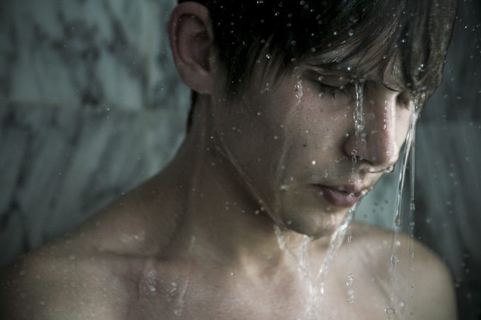 Shirtless young man with wet hair stands under shower water with eyes closed, appearing introspective against a marble background.
