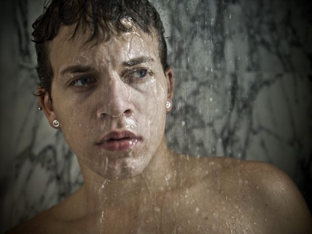 Shirtless young man with wet hair and earrings stands under running water in a marble shower, looking contemplative.