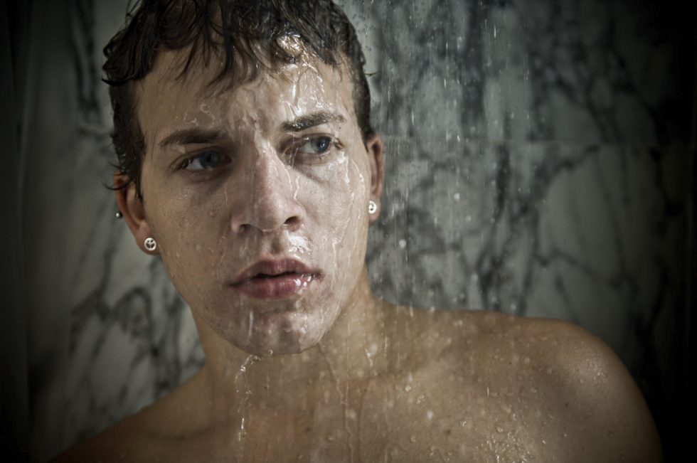 Shirtless young man with wet hair and earrings stands under running water in a marble shower, looking contemplative.