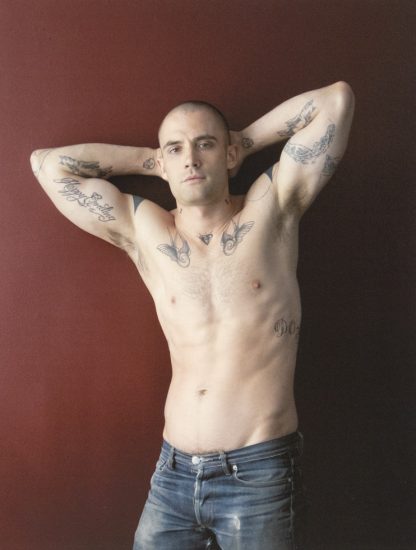 Shirtless man with tattoos poses against a red wall, arms behind head, wearing low-rise jeans.