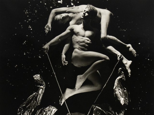 A black and white photograph of two nude men in a black abyss with splashes of light in the darkness.