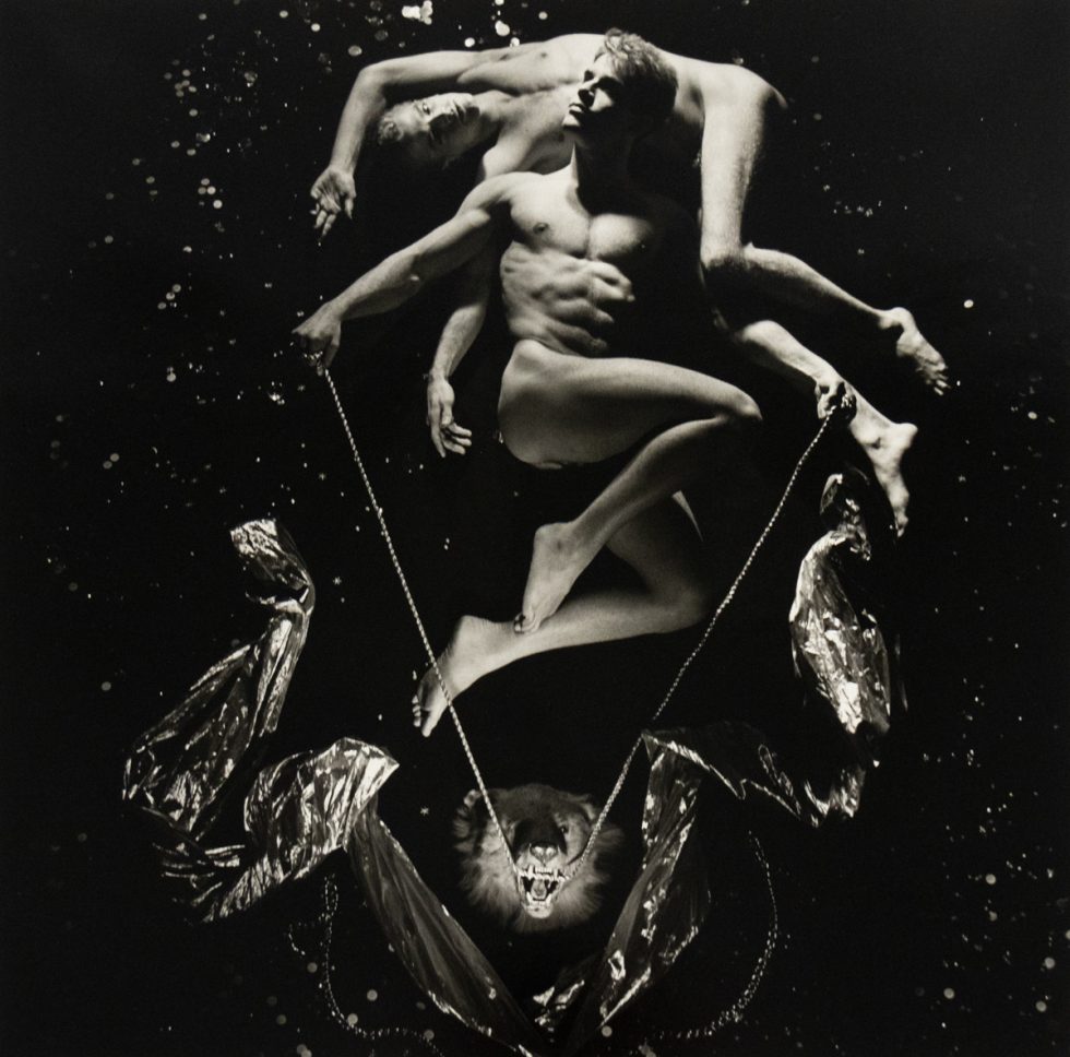 A black and white photograph of two nude men in a black abyss with splashes of light in the darkness.