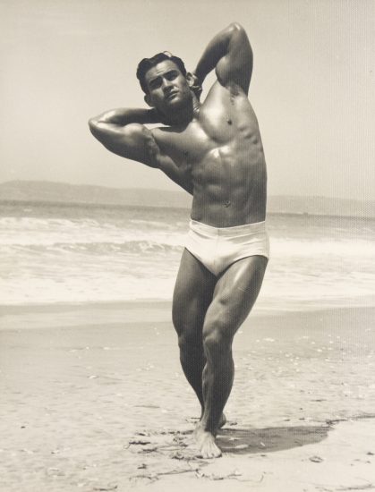 Muscular man in white trunks stands barefoot on a beach boardwalk, gazing downward in dramatic sunlight.
