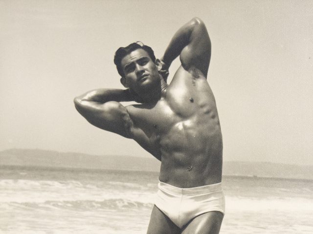 Muscular man in white trunks stands barefoot on a beach boardwalk, gazing downward in dramatic sunlight.