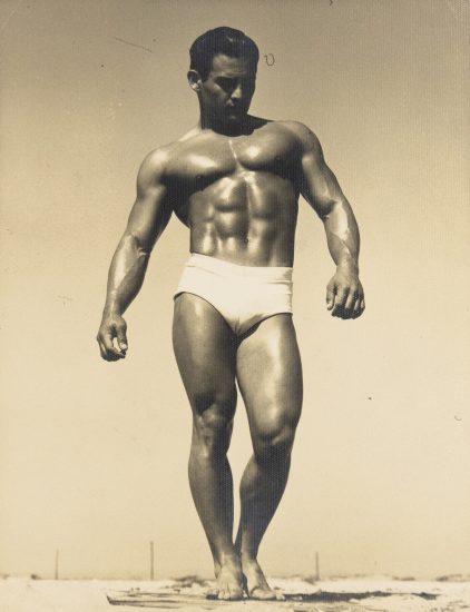 Muscular man in white trunks stands barefoot on a beach boardwalk, gazing downward in dramatic sunlight.