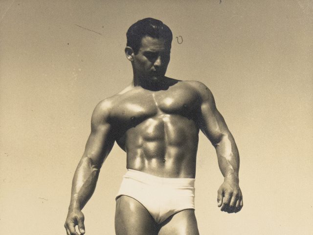 Muscular man in white trunks stands barefoot on a beach boardwalk, gazing downward in dramatic sunlight.