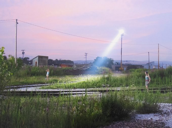 Two girls stand apart in overgrown train tracks as a bright spotlight cuts through dusk.