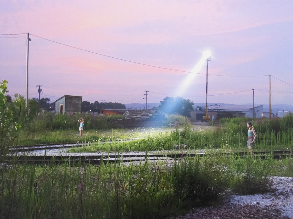 Two girls stand apart in overgrown train tracks as a bright spotlight cuts through dusk.