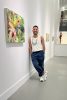 A color photo of a man leaning against a wall with paintings on display.