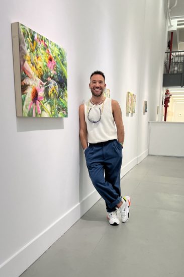 A color photograph of a man leaning against a wall with paintings on display.