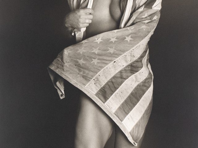 Shirtless man partially wrapped in a worn American flag, revealing muscular legs and torso against a dark background.