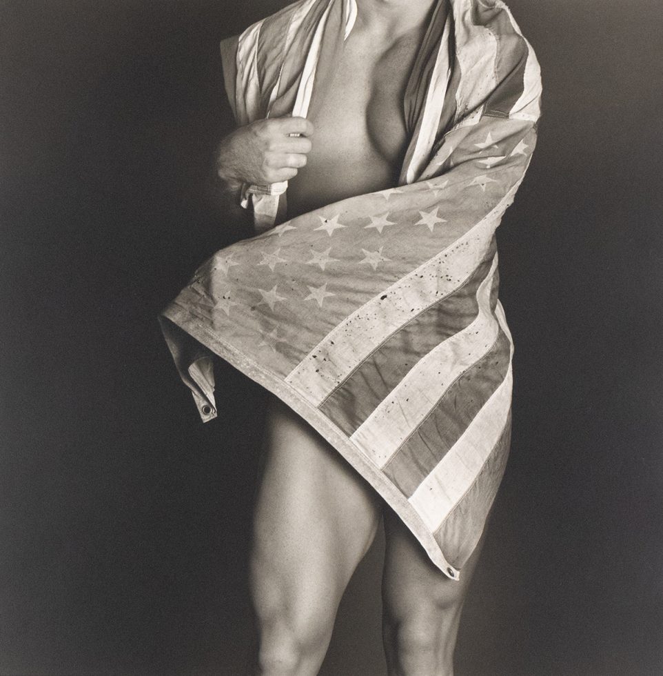 Shirtless man partially wrapped in a worn American flag, revealing muscular legs and torso against a dark background.