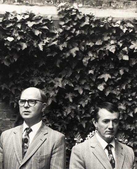 Click for sizes / editions / prices. Two men in matching striped suits and ties stand side by side in front of a dense wall of ivy, facing forward with serious expressions.