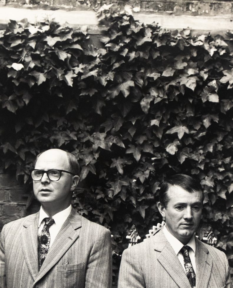 Two men in matching striped suits and ties stand side by side in front of a dense wall of ivy, facing forward with serious expressions.