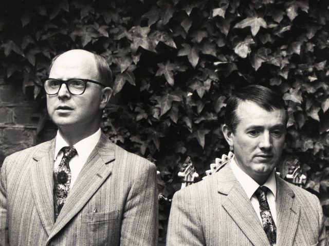 Two men in matching striped suits and ties stand side by side in front of a dense wall of ivy, facing forward with serious expressions.