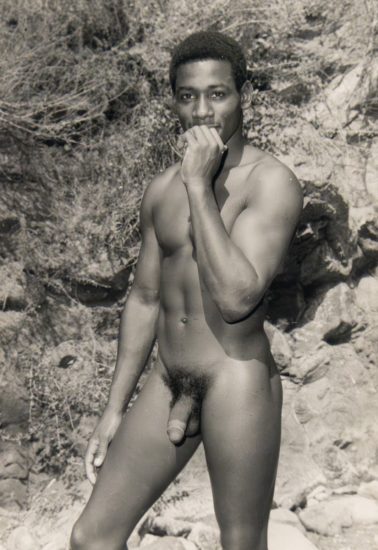 Black-and-white photo of a nude man standing outdoors, holding one hand to his mouth and gazing directly at the camera.