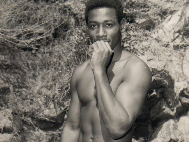 Black-and-white photo of a nude man standing outdoors, holding one hand to his mouth and gazing directly at the camera.