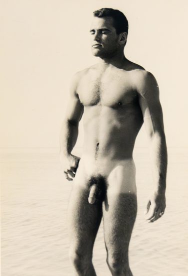 A black and white photograph of a nude man with prominent tan lines.