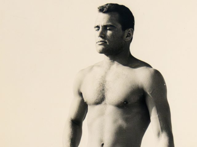 A black and white photograph of a nude man with prominent tan lines.