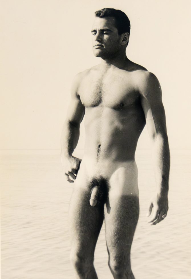 A black and white photograph of a nude man with prominent tan lines.