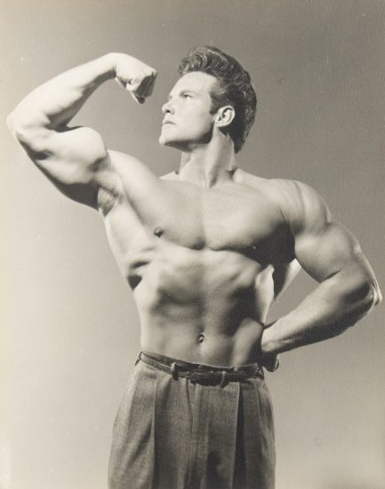 Shirtless male bodybuilder flexing his arm in classic pose, wearing high-waisted trousers in a vintage black-and-white photo.