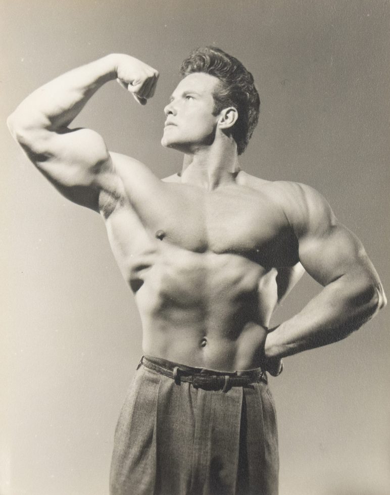 Shirtless male bodybuilder flexing his arm in classic pose, wearing high-waisted trousers in a vintage black-and-white photo.