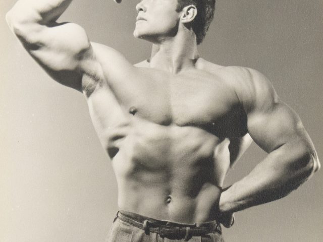 Shirtless male bodybuilder flexing his arm in classic pose, wearing high-waisted trousers in a vintage black-and-white photo.