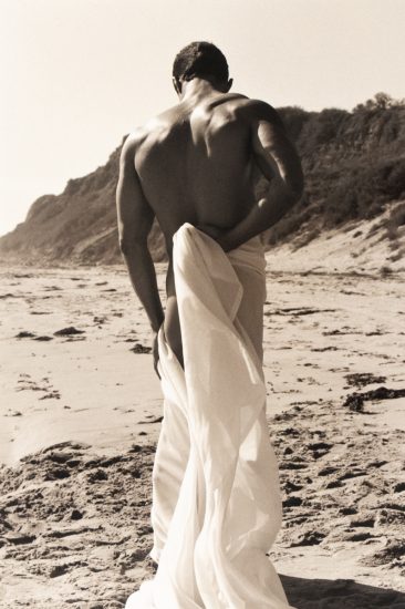 Shirtless man with muscular back stands on beach draped in flowing white fabric, facing away, with cliffs in the background.