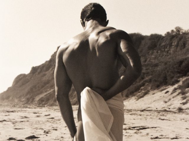 Shirtless man with muscular back stands on beach draped in flowing white fabric, facing away, with cliffs in the background.