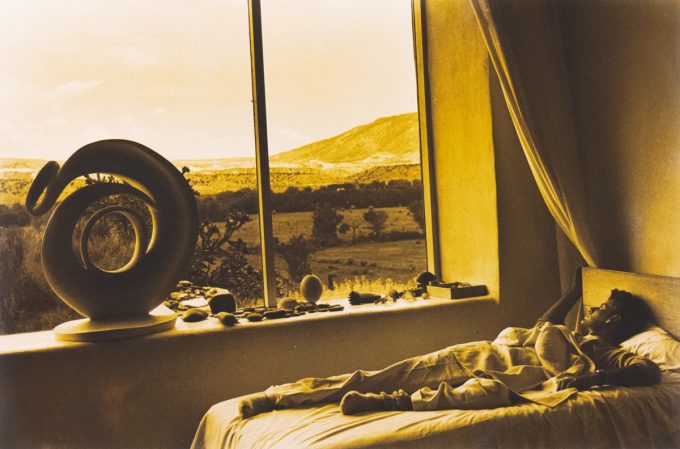 Man lies in bed beside a large window overlooking a desert landscape, with stones and a spiral sculpture on the windowsill.