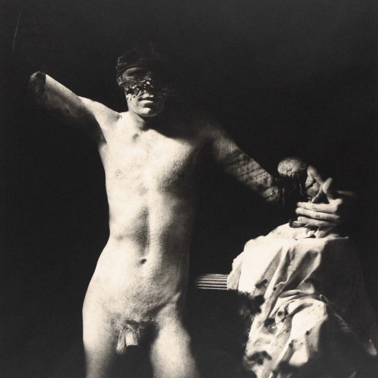 A disfigured, nude male figure, resembling a classical statue, is dramatically lit against a dark background in this black and white photograph.