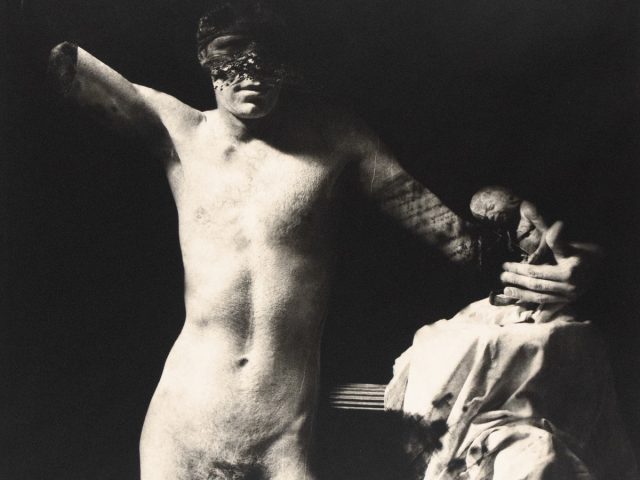 A disfigured, nude male figure, resembling a classical statue, is dramatically lit against a dark background in this black and white photograph.