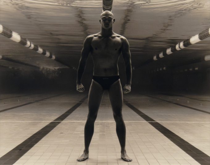 Underwater figure stands powerfully at the bottom of an empty pool, framed by lane lines and dramatic light, with fists clenched and legs apart.