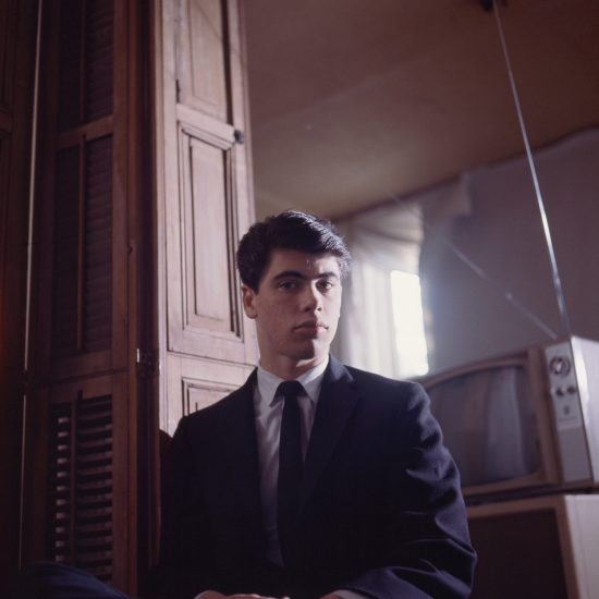 A serious young person with dark hair and a suit looks directly at the camera, with wooden paneling and a vintage TV in the background.