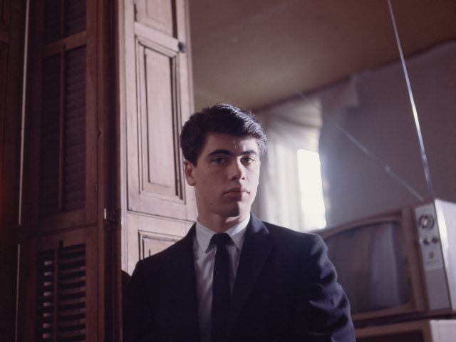 A serious young person with dark hair and a suit looks directly at the camera, with wooden paneling and a vintage TV in the background.