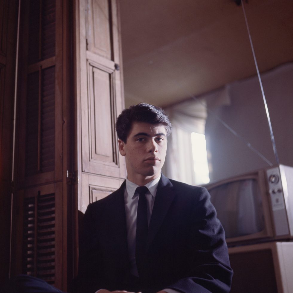 A serious young person with dark hair and a suit looks directly at the camera, with wooden paneling and a vintage TV in the background.