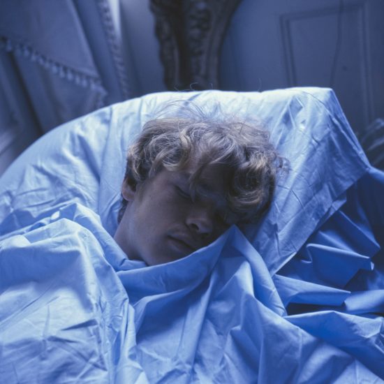 A fair-skinned person with curly blond hair sleeps soundly, covered in light blue sheets and illuminated by a cool, blue light.
