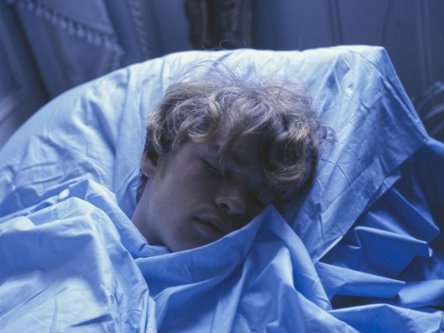 A fair-skinned person with curly blond hair sleeps soundly, covered in light blue sheets and illuminated by a cool, blue light.
