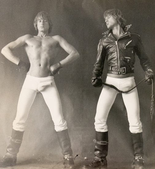 A black and white photograph of two men in white tights, one in a leather jacket.