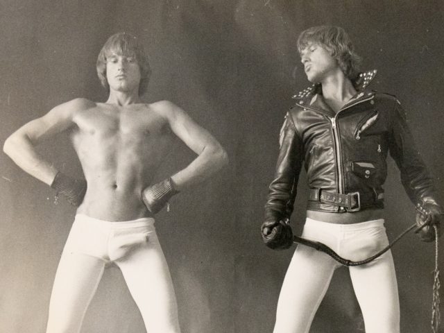 A black and white photograph of two men in white tights, one in a leather jacket.