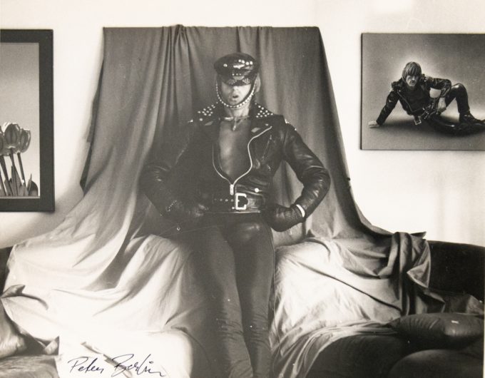 A man in tight leather pants and a studded jacket poses confidently in front of draped fabric, flanked by framed photos of himself.