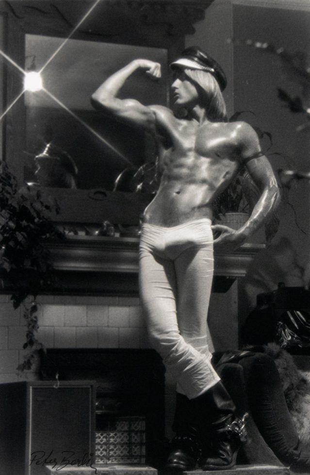 Shirtless person in tight pants and boots strikes a flexing pose indoors, standing in front of a mirror above a fireplace in soft, dramatic lighting.