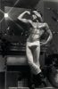 Shirtless person in tight pants and boots strikes a flexing pose indoors, standing in front of a mirror above a fireplace in soft, dramatic lighting.