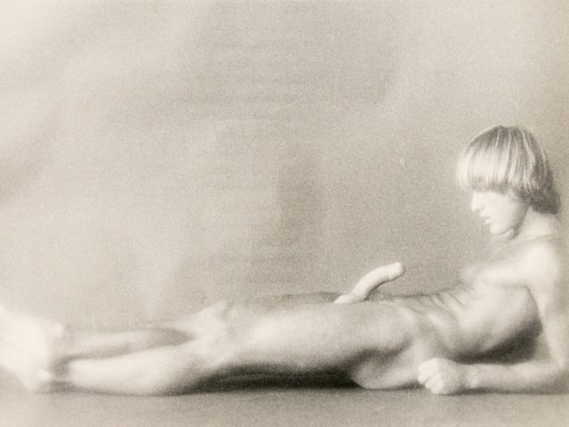 A nude figure reclines on the floor, gazing downward, in a softly lit and blurred black-and-white composition.